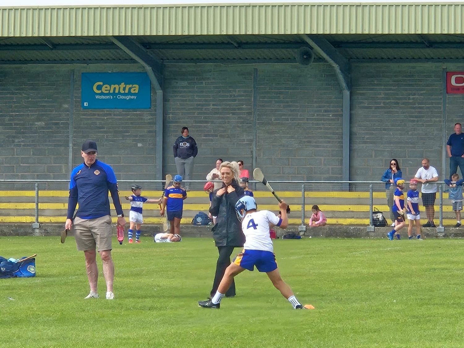 A young hurler striking the ball during a match while a coach looks on