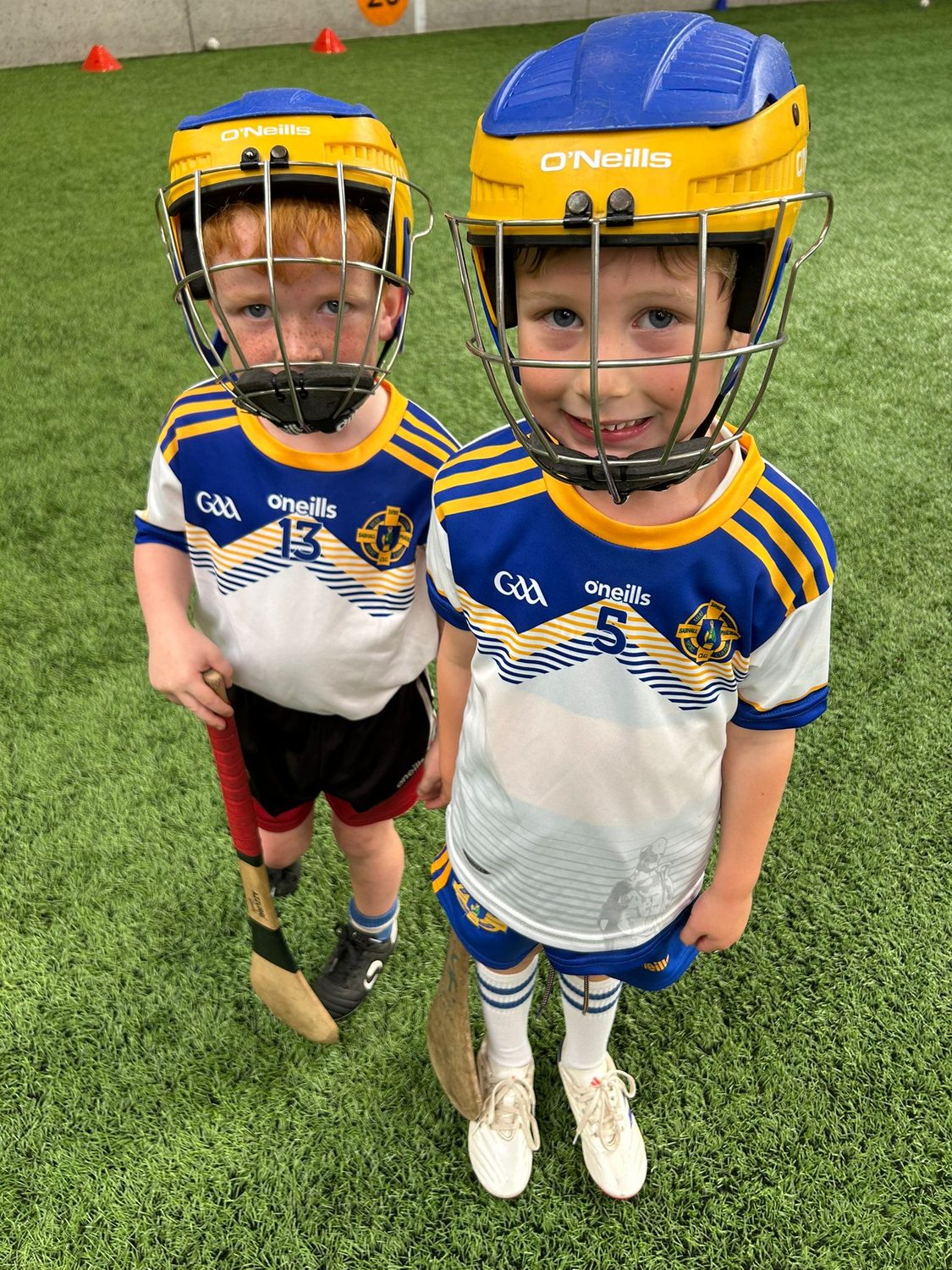 Two young hurlers in helmets and club jerseys on the indoor 3G pitch, hurls in hand